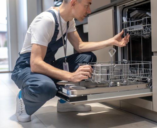 Diagnostics of equipment. Profile of serious man in dark overalls with badge crouching attentively looking at open dishwasher in kitchen at home during day